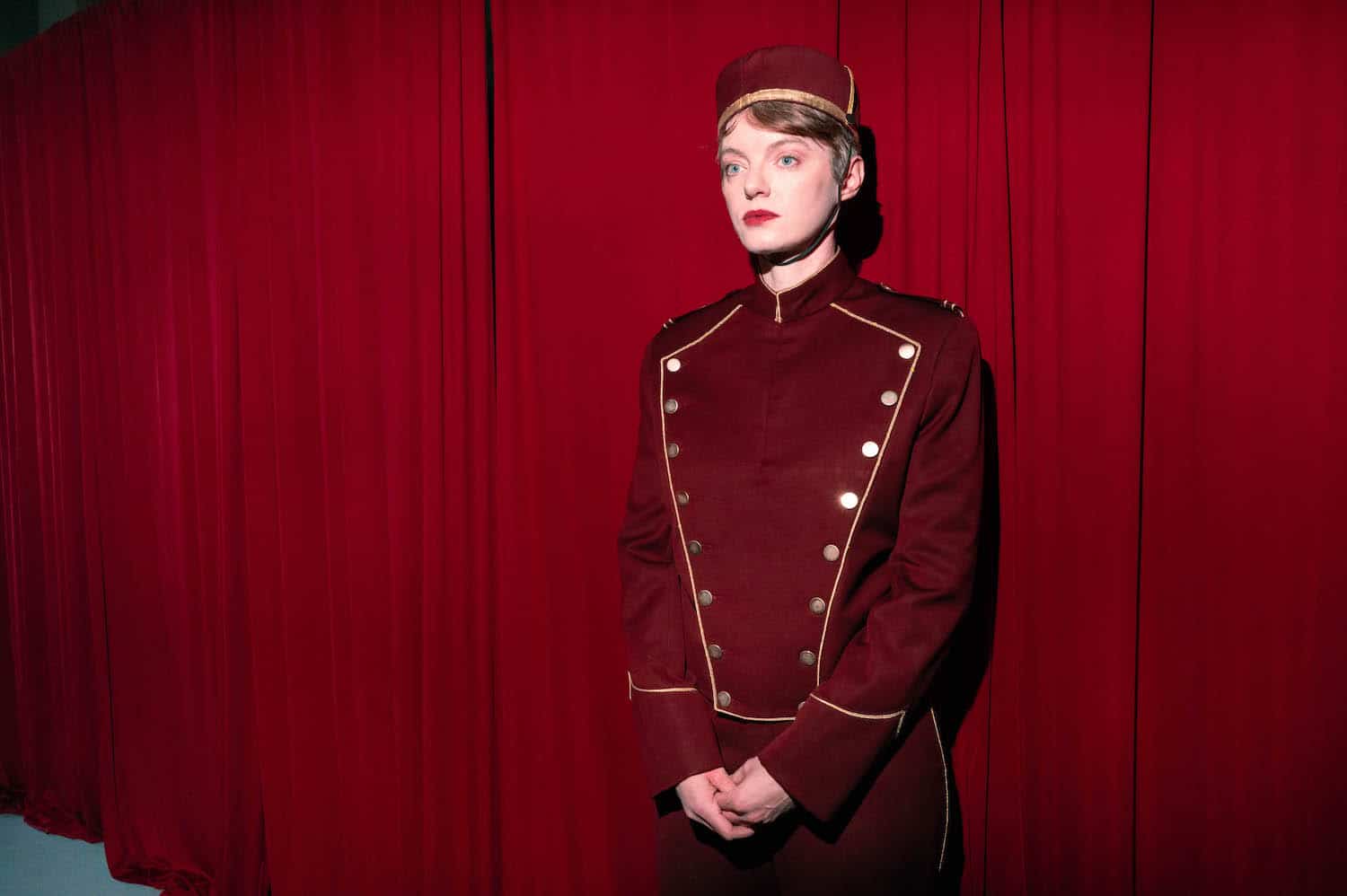 An usher in a red bellhop uniform standing against the red curtains